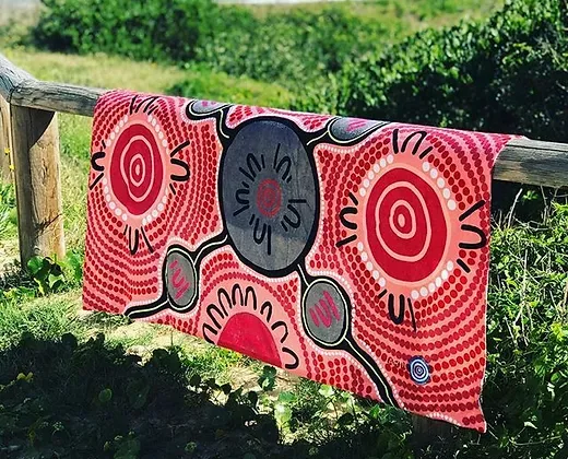 Colorful Aboriginal-inspired towel with circular patterns draped over a rustic wooden fence in a grassy area.