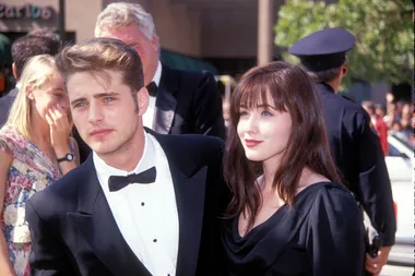 A man and woman in formal attire at an outdoor event, surrounded by people and a police officer in the background.