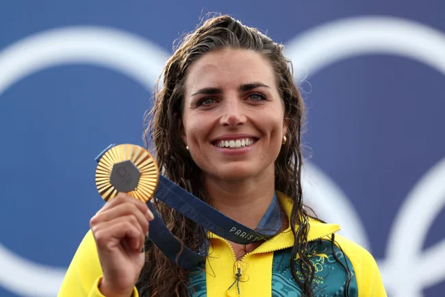 Woman in yellow jacket smiles, holding a gold medal. Olympic rings in the background.