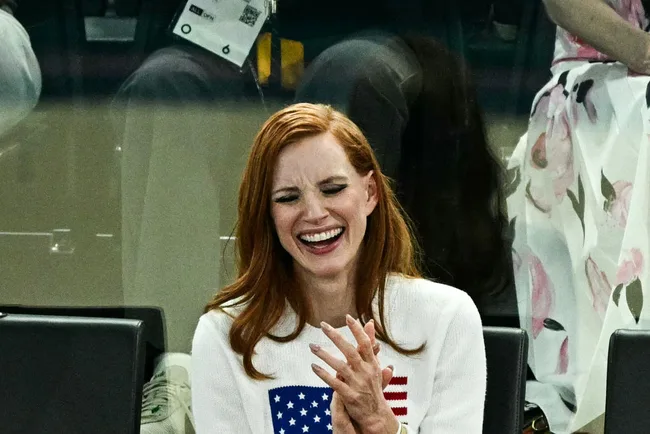 Red-haired woman in a white sweater with a flag design, laughing joyfully at an indoor event.