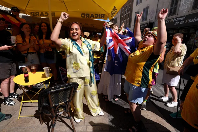 A joyful group of Australian sports fans in yellow outfits celebrate outdoors, holding an Australian flag.
