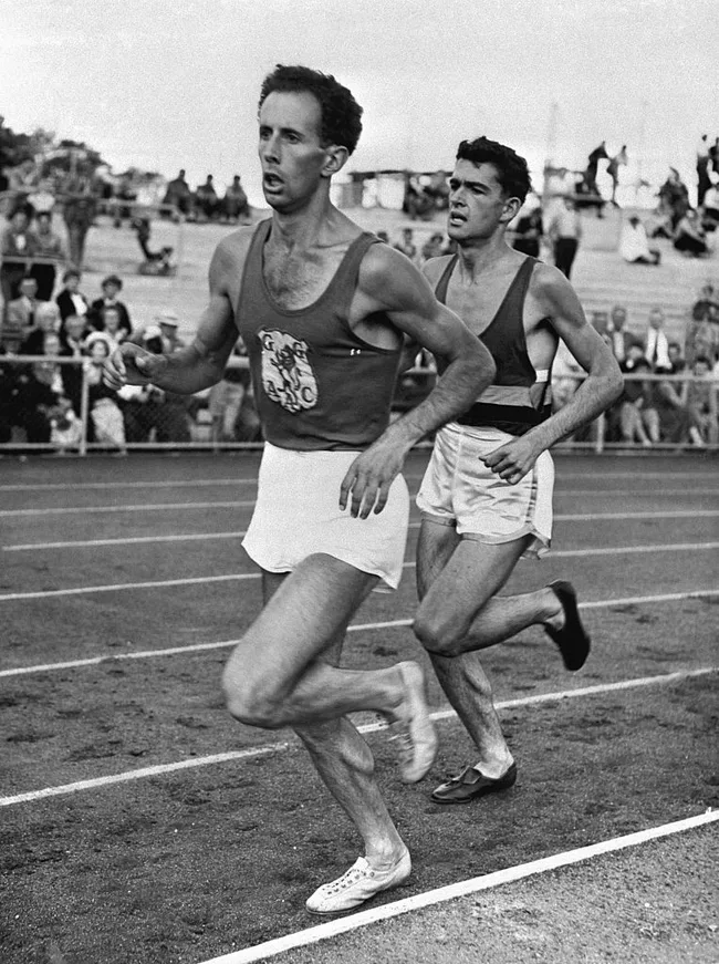 Two male athletes running on a track during a race, with a crowd watching in the background.