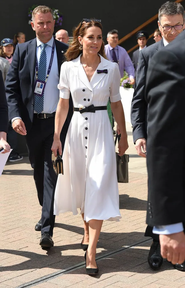 A woman in a white dress with black buttons walks outdoors, accompanied by security personnel and others.