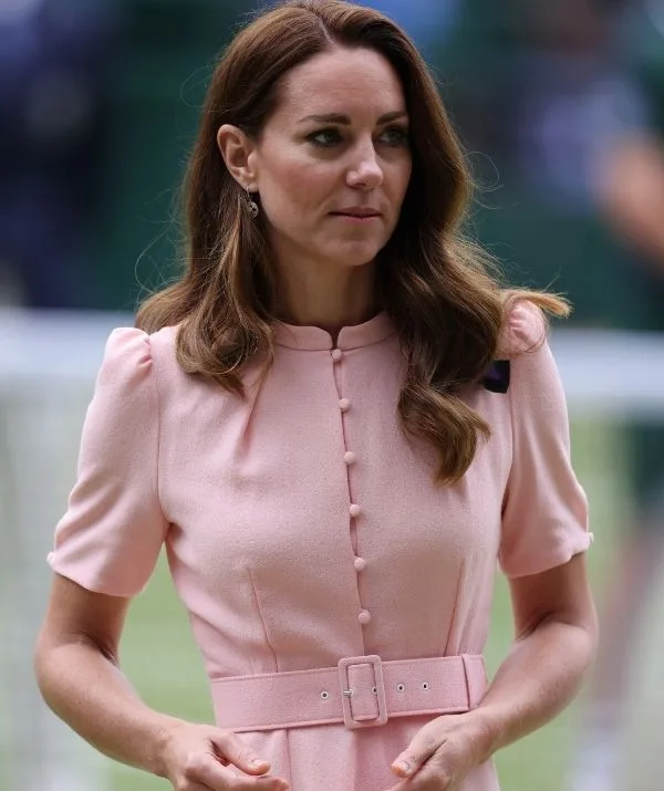 A woman with long brown hair in a pink dress with a belt stands outdoors on a blurred green background.