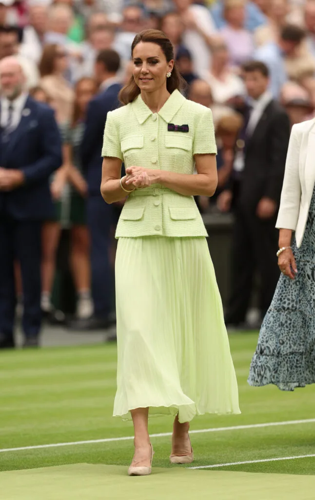 A woman in a light green suit walks on a grassy field at a formal event, surrounded by a crowd.