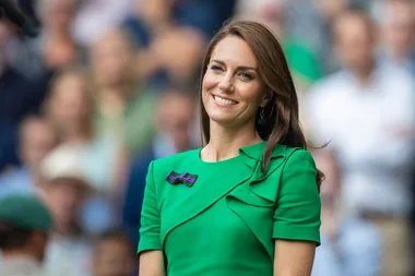 A woman in a green dress with a smile, wearing a bow pin and green earrings, stands in front of a blurred crowd.