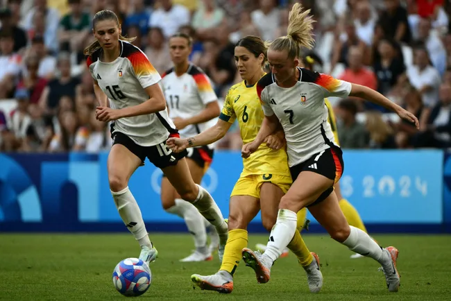 Soccer players in white and yellow jerseys compete in a match, with a colorful ball on a green field, surrounded by spectators.