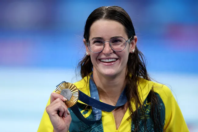 A female athlete wearing glasses and a yellow jacket smiles while holding a gold medal.