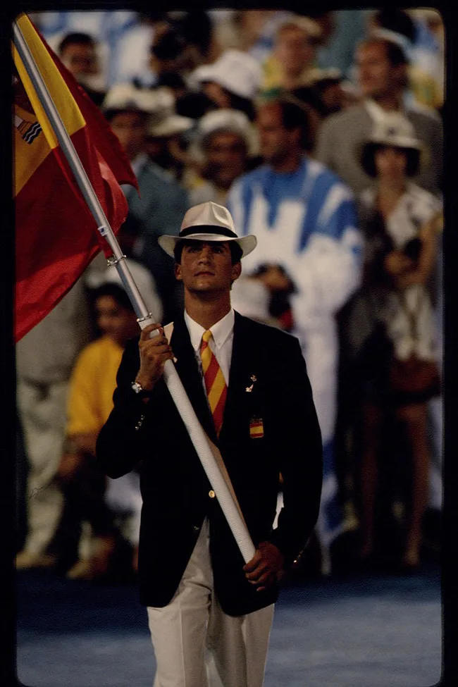 A person in a suit and hat carries the Spanish flag at an Olympic event, surrounded by a crowd.