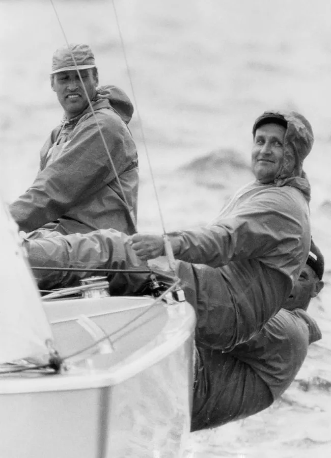 Two men in rain gear sailing a small boat, one smiling and the other focused, with a third person partially visible.