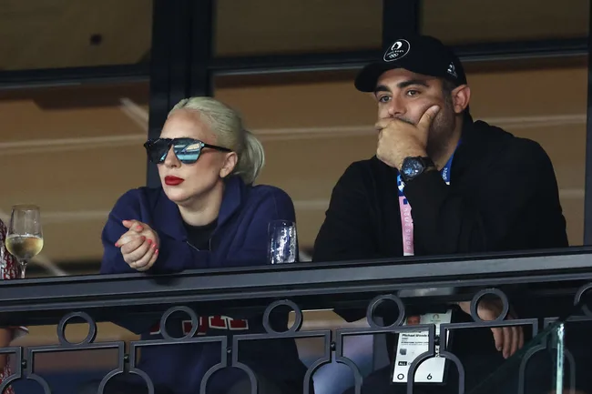 Two people watching intently from a balcony, one wearing sunglasses and a blue jacket, the other in a black cap.