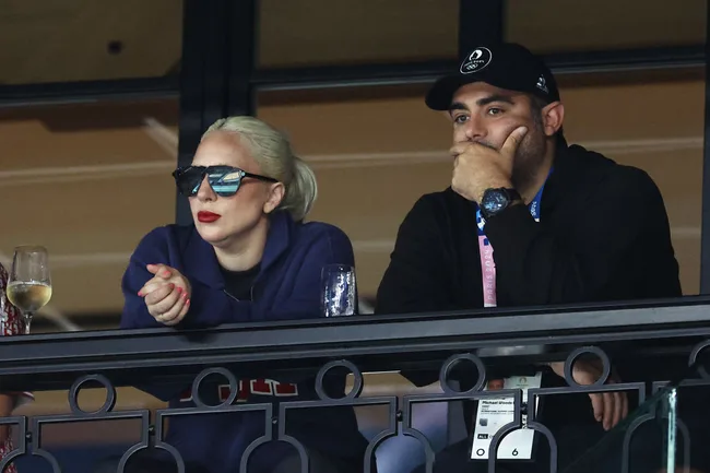 Two people watching intently from a balcony, one wearing sunglasses and a blue jacket, the other in a black cap.