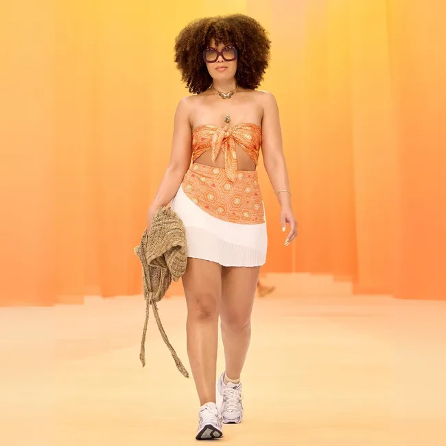 A woman in a colorful top and skirt walks confidently on a runway, wearing sunglasses and holding a bag.