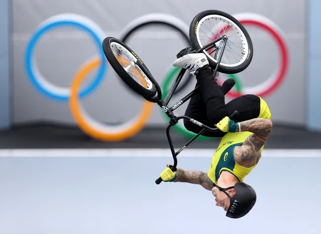 A cyclist performs a BMX trick upside down against Olympic rings backdrop.
