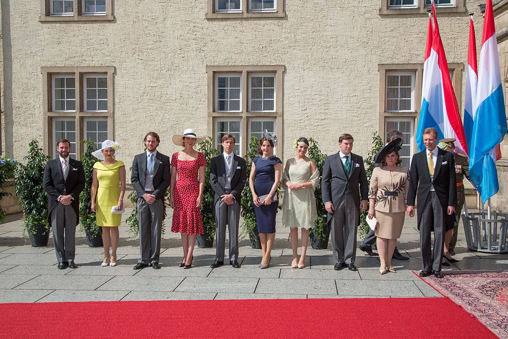 A group of ten people in formal attire standing on a red carpet with Luxembourg flags in the background.
