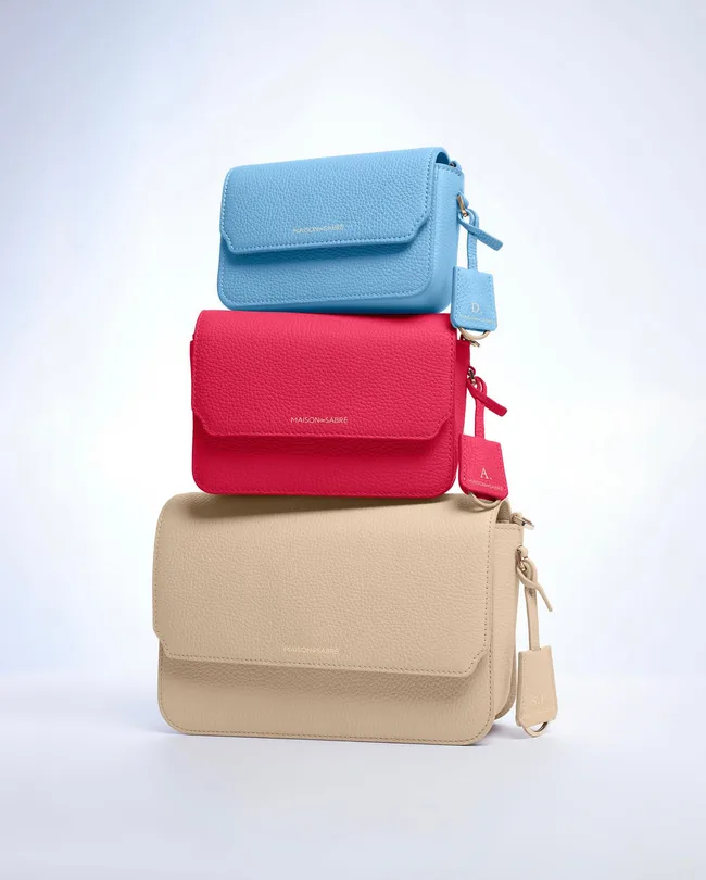 Three Maison de Sabre leather bags stacked: blue on top, red in the middle, and beige at the bottom, with monogram tags.
