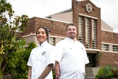 Two chefs in white jackets stand outside a building with a MasterChef logo.