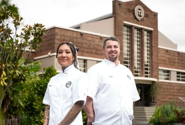 Two chefs in white jackets stand outside a building with a MasterChef logo.