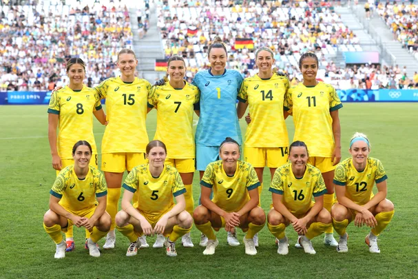 Australia women's soccer team in yellow jerseys, posing on field during a match, with fans in background.