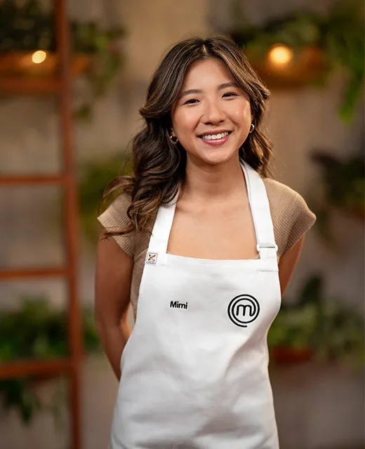 Smiling person named Mimi in a MasterChef apron, standing in a kitchen setting with plants and warm lighting.