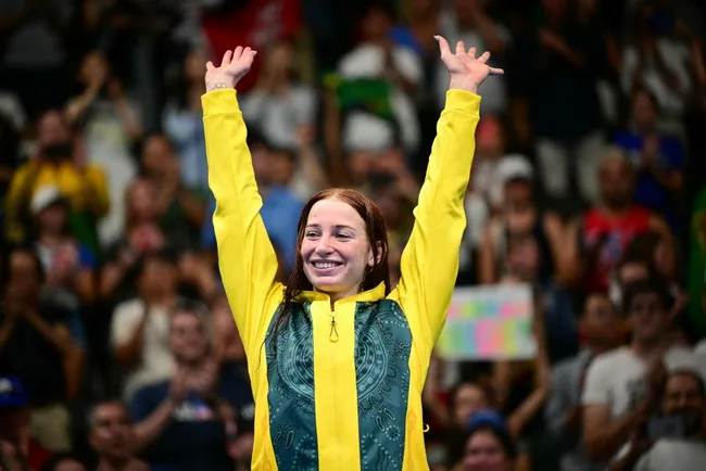 A woman in a yellow jacket raises her arms in celebration at a sports event, with a cheering crowd in the background.