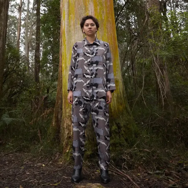 Person in camo outfit standing against a large tree in a forest setting.