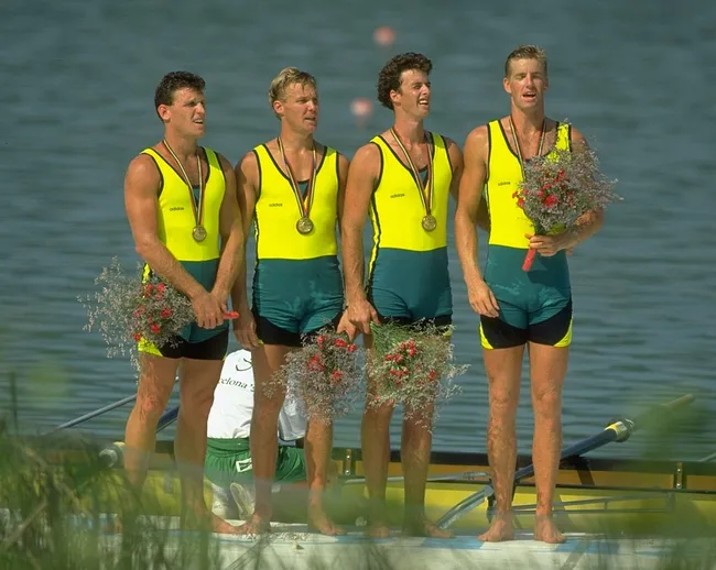 Four male rowers in Australia uniforms hold medals and flowers at a podium with water in the background.