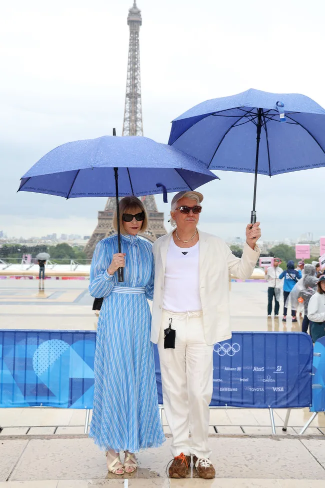 Two people stand in front of the Eiffel Tower holding blue umbrellas on a rainy day.