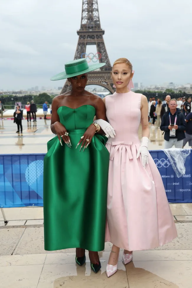 Two individuals in elegant green and pink dresses pose near the Eiffel Tower, with the Olympic rings in the background.