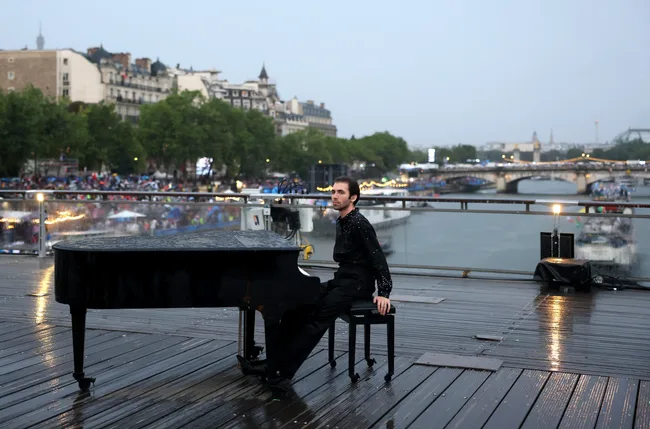 Person seated at a grand piano on a rainy deck overlooking a river and cityscape, with a bridge and buildings in background.