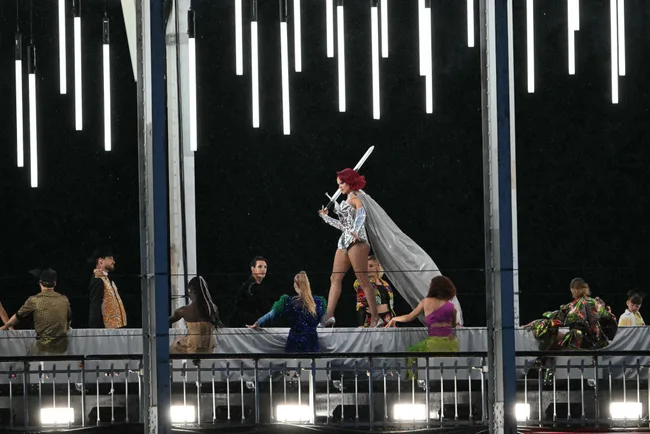 Performer in silver costume with red hair holding sword on stage with dancers under vertical lights.