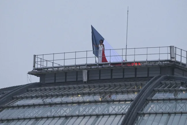Person in a white outfit stands on a rooftop holding a large French flag.