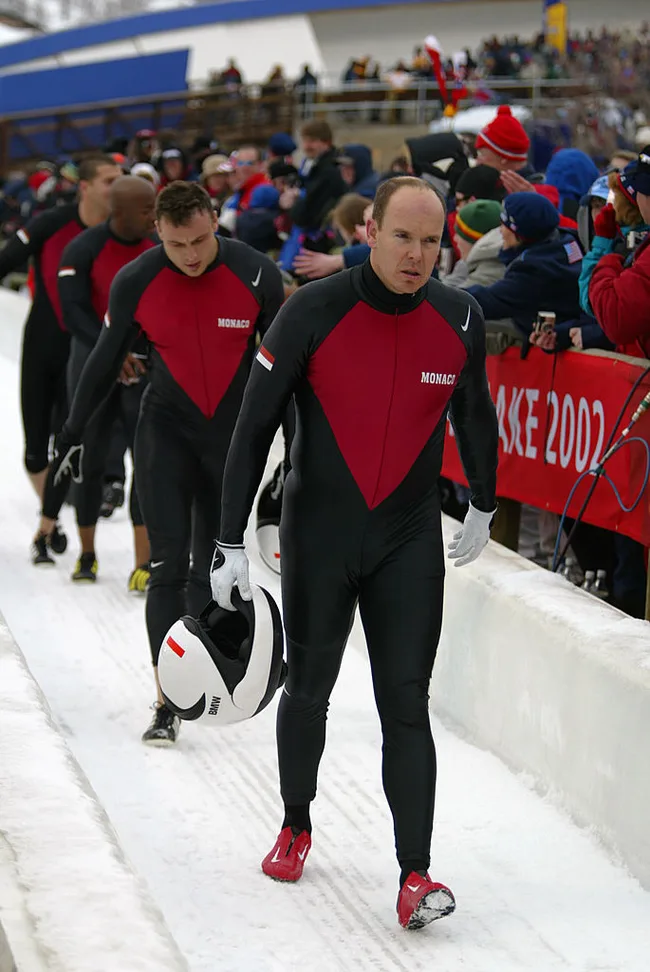 Monaco bobsled team walks past spectators at the 2002 Winter Olympics, holding helmets, in black and red suits.