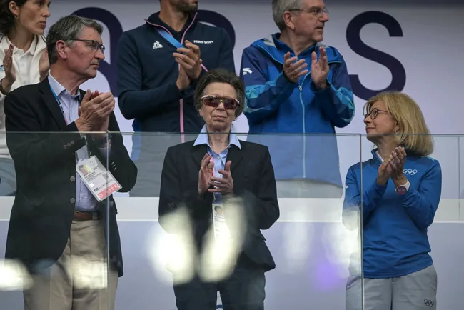 Group of people clapping at an event, some wearing official badges and Olympic-themed attire, standing behind a glass barrier.