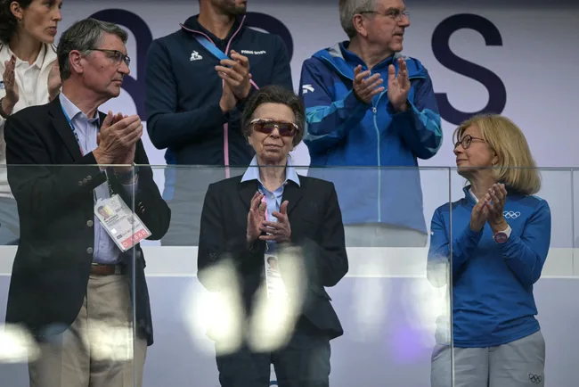 Group of people clapping at an event, some wearing official badges and Olympic-themed attire, standing behind a glass barrier.