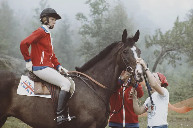 Rider in red jacket on a horse with Union Jack saddlecloth, assisted by two people, outdoors.