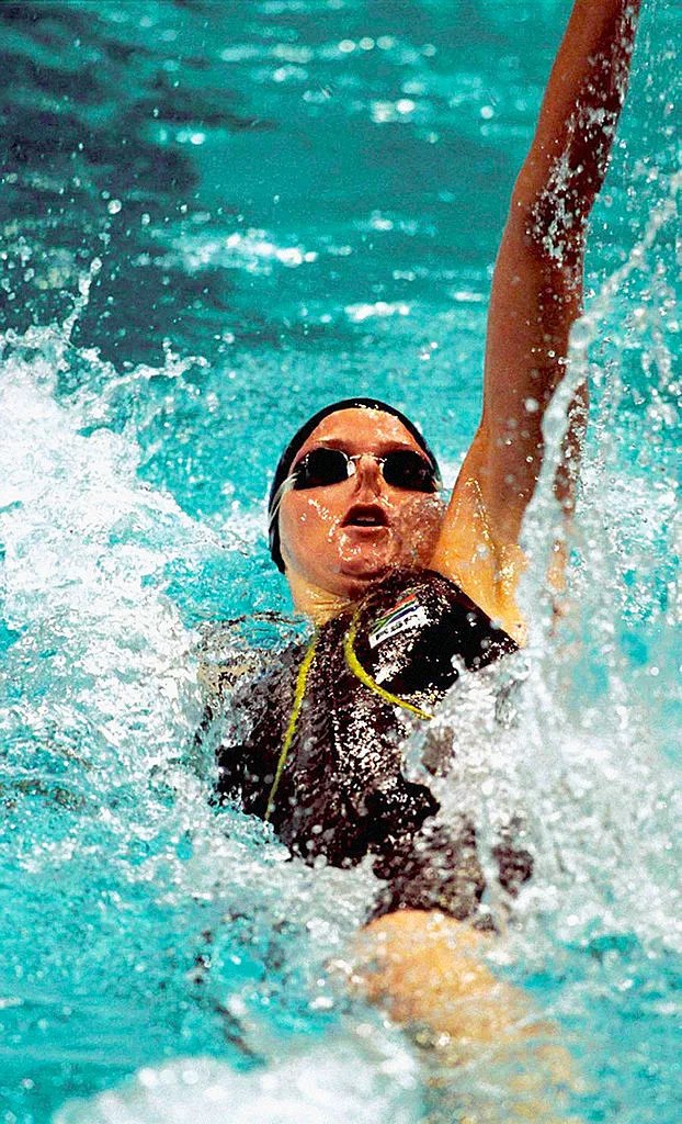 A swimmer wearing goggles and a swim cap participates in a backstroke event, creating splashes in the pool.