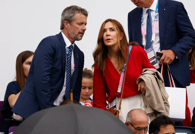 A man in a blue suit and a woman in a red blouse talking in a sports event crowd.