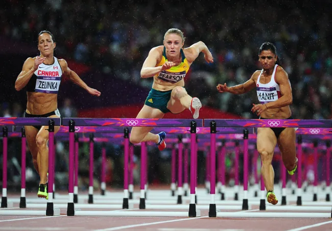 Three athletes in a hurdling race at the London 2012 Olympics, with rain visible in the background.