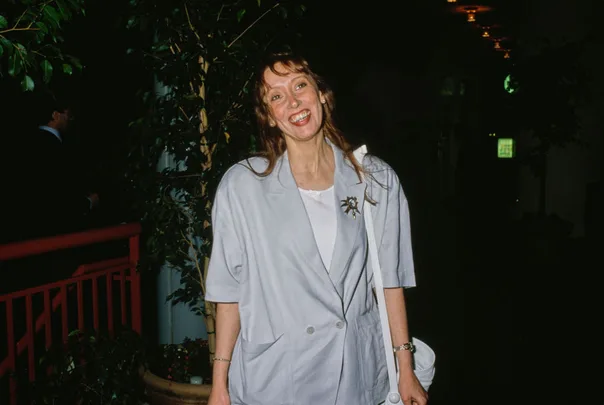 A woman smiling in a light blue blazer stands indoors near leafy plants.
