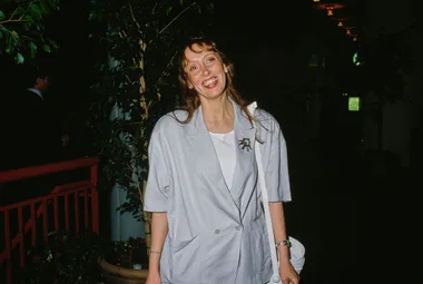 A woman smiling in a light blue blazer stands indoors near leafy plants.