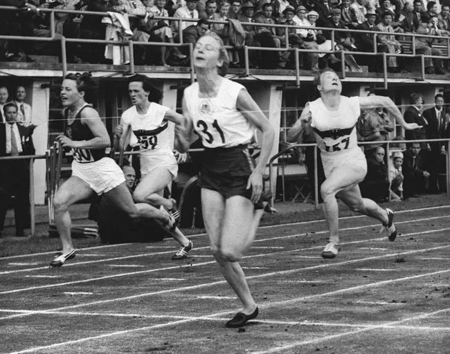 Women athletes sprinting towards the finish line in a race, with a crowded grandstand in the background.