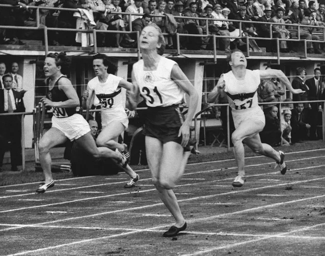 Women athletes sprinting towards the finish line in a race, with a crowded grandstand in the background.