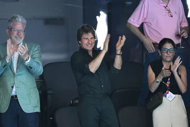 Three people clapping in a stadium luxury box, one wearing a green jacket, another in black shirt, and a woman with glasses.