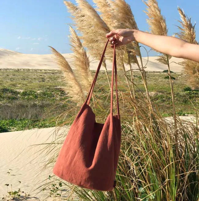 Hand holding a red tote bag against a background of pampas grass and sand dunes under a clear blue sky.