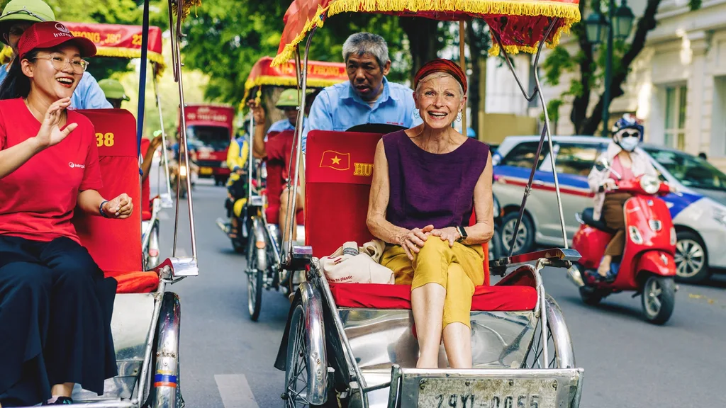 Three people ride rickshaws on a city street, smiling. A woman in red drives one, and a woman in purple rides another.
