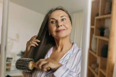 Beautiful, charming elderly female combing grey hair with round hairbrush standing against shelves in hallway or bedroom in front of mirror, looking at her reflection, getting ready to go to bed.