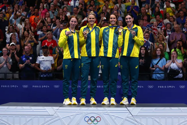 Australian women's sports team on podium with gold medals at Paris 2024 Olympics, smiling with cheering crowd.