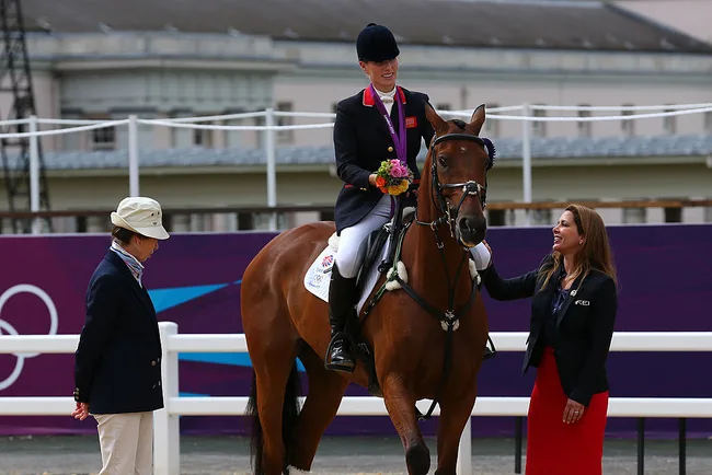 Equestrian champion on a horse holding flowers, flanked by two women, with an Olympic backdrop.