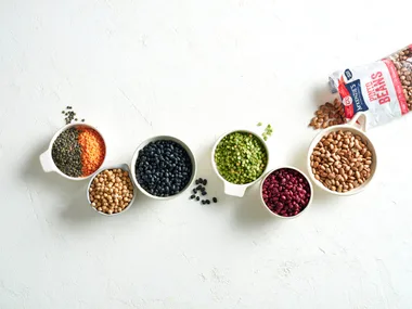 Assorted beans and lentils in bowls on a white surface, with a McKenzie's Pinto Beans package in the background.